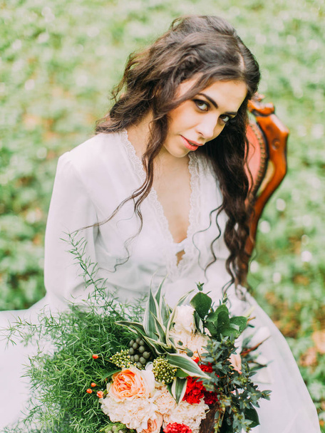 The lovely close-up portrait of the woman in the white dress, sitting on the chair, holding the beautiful bouquet of flowers and looking at the camera. Forest location. - Foto, Imagen
