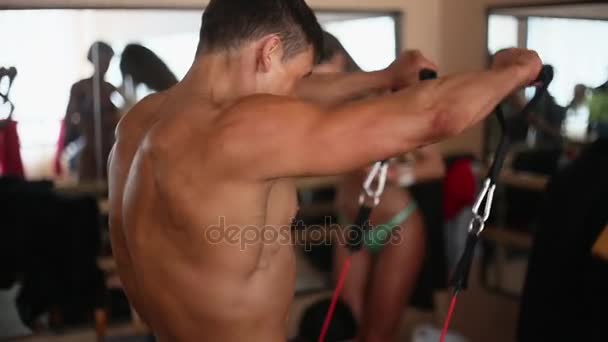 Vista laterale, posteriore del bodybuilder maschile con busto nudo che fa esercizio con banda di resistenza. Donna in bikini sullo sfondo
 - Filmati, video