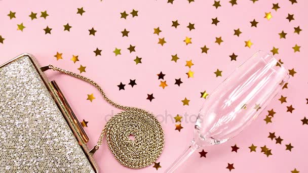 Festive evening golden clutch and champagne glass with star sprinkles shining on pink. Holiday and celebration background. Luxury accessories and party concept - Footage, Video