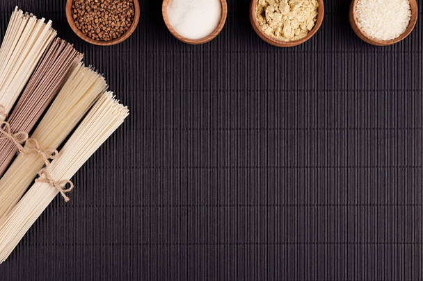 Decorative border of bundles raw noodles with ingredient in wooden bowls on black striped mat background with copy space, top view. - Photo, image