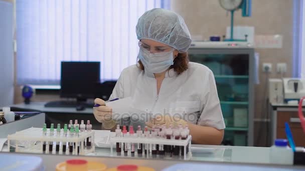 Výzkumný asistent v bílém laboratorním drhne, ochranné brýle, masky, čepici a rukavice sedí u stolu v lékařské laboratoři se sadou zkumavek a příprava vzorků krve k vyšetření. - Záběry, video