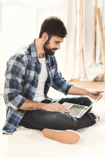 Hombre sentado en la alfombra y trabajando en el ordenador portátil
 - Foto, Imagen