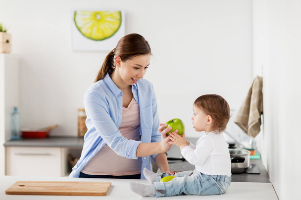 mother giving green apple to baby at home kitchen - Фото, изображение