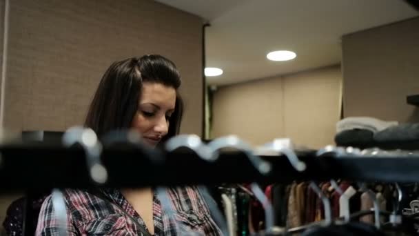 Μελαχρινή γυναίκα σε καρώ πουκάμισο έρχεται σε rack με κρεμάστρες για κατάστημα - Πλάνα, βίντεο