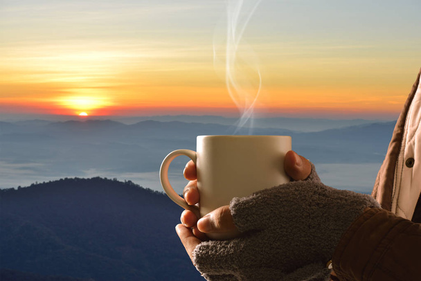 Τα χέρια με γάντια κρατώντας φλιτζάνι καφέ στο βουνό και τον ήλιο φόντο - Φωτογραφία, εικόνα