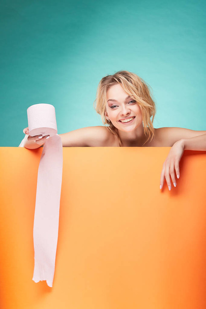 Young beautiful blonde woman holding roll of toilet paper with smiling face in studio on turquoise background and orange sheet  - Фото, изображение