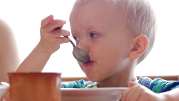Baby 2 years old eats porridge with a spoon. Mom sits next to the boy. home furnishings - Felvétel, videó