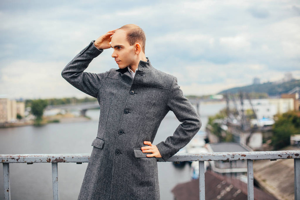 portrait de jeune homme debout sur le pont et posant mode vogue, temps nuageux et froid venteux, arrière-plan ville, détournant les yeux. dans un long manteau gris élégant
 - Photo, image