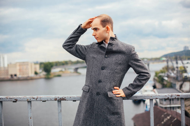 muotokuva nuori mies seisoo sillalla ja poseeraa muoti muodissa, pilvinen ja kylmä tuulinen sää, tausta kaupunki, katselee pois. pitkä harmaa takki tyylikäs
 - Valokuva, kuva