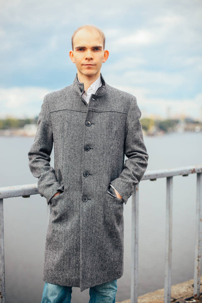 retrato de un joven de pie en el puente y posando moda de moda, nublado y frío viento, ciudad de fondo, mirando hacia otro lado. en un largo abrigo gris elegante
 - Foto, imagen