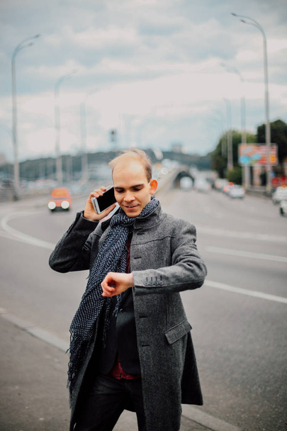 portrait of young man standing near the road with heavy traffic and posing, cloudy and cold windy weather closeup, talking on the phone with business partners, the wind blows long stylish gray coat - Fotó, kép