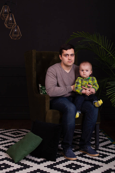 Dad and his son sitting on green chair with a bright colorful pillow with palm tree near them. modern, minimalist. dark interior. hugging with toddler. Child making funny facial expressions, smiling - Fotografie, Obrázek