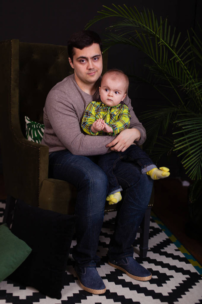 Papa et son fils assis sur une chaise verte avec un oreiller coloré lumineux avec un palmier près d'eux. moderne, minimaliste. intérieur sombre. câlin avec tout-petit. Enfant faisant des expressions faciales drôles, souriant
 - Photo, image