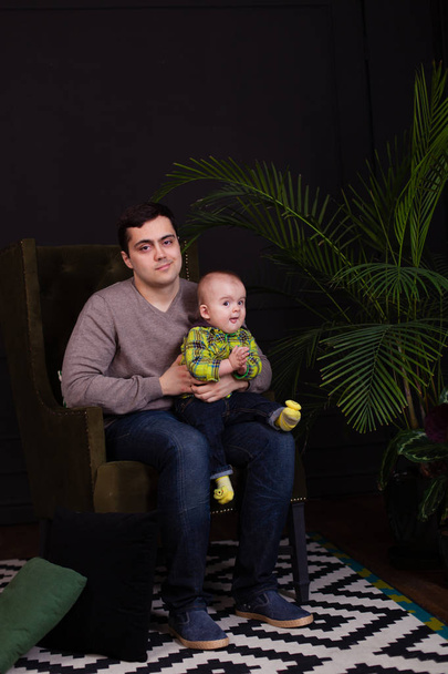 Papa et son fils assis sur une chaise verte avec un oreiller coloré lumineux avec un palmier près d'eux. moderne, minimaliste. intérieur sombre. câlin avec tout-petit. Enfant faisant des expressions faciales drôles, souriant
 - Photo, image
