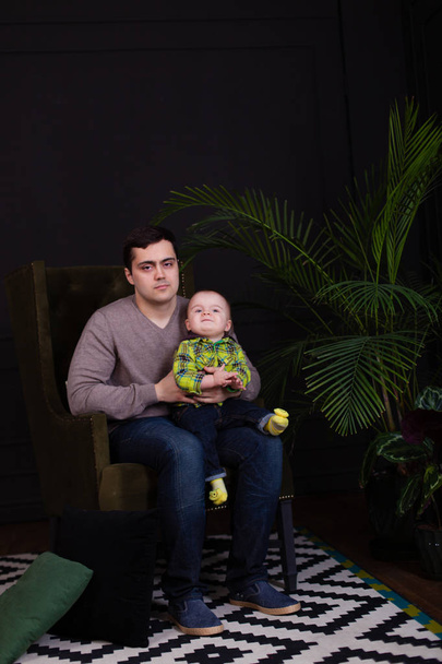 Tata i syn siedzi na krześle zielony z jasny kolorowy poduszkę z palmą w ich pobliżu. nowoczesne, minimalistyczne. Mroczne wnętrza. przytulanie się z malucha. Dziecko co zabawny wyraz twarzy, uśmiechając się - Zdjęcie, obraz