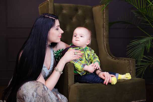 Mooie jonge Kaukasische moeder die haar baby-zoontje in haar armen in de stijlvolle studio op een achtergrond van gloeilampen in een modernistische stijl, knuffelen haar zoon. minimalisme, interieur, lage sleutel - Foto, afbeelding