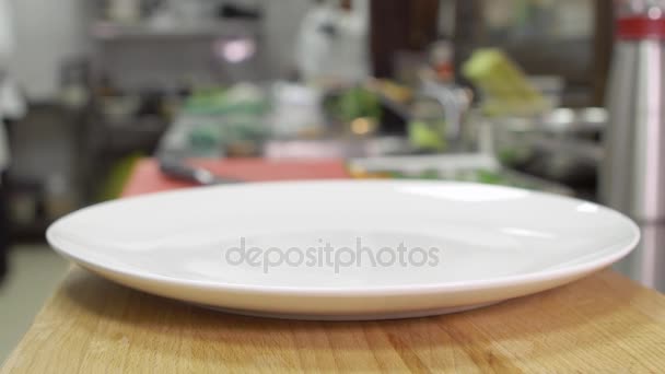 Na dřevěný stůl stojí prázdný čistý bílý talíř. Deska připravena sloužit jídlo v restauraci nebo kavárně. V pozadí typické kuchyňka - Záběry, video
