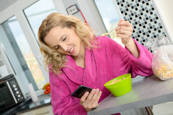 Młoda blond kobieta jedzenie zbóż i telefon w godzinach porannych - Zdjęcie, obraz