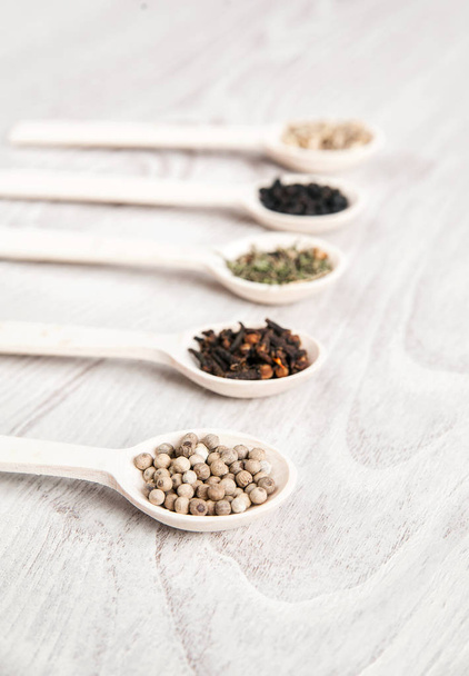 Various spices and herbs in wooden spoons on white table background. Aromatic food cooking ingredients. Black and white pepper, clove, savory, fennel seeds. - Фото, зображення