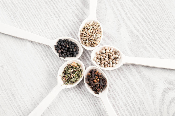 Various spices and herbs in wooden spoons on white table background. Black and white pepper, clove, savory, fennel seeds. - Foto, afbeelding