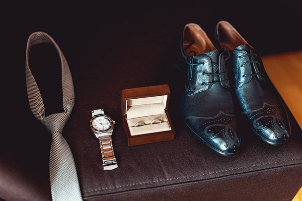 Primer plano de los accesorios modernos del novio. anillos de boda en una caja de madera marrón, corbata, zapatos de cuero y reloj
 - Foto, imagen
