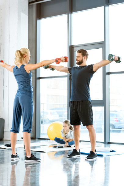 Fitnessmenschen trainieren mit Kurzhanteln, während Teenager-Mädchen im Fitnessstudio auf Matte sitzen - Foto, Bild