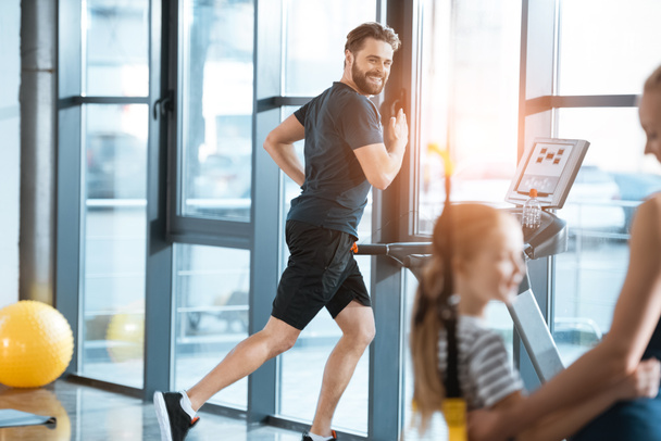 Woman with girl looking at handsome man workout on treadmill at gym - Photo, Image