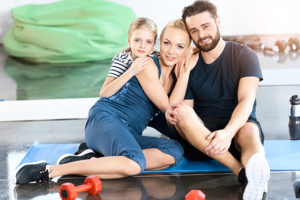 Портрет счастливой семьи сидящей на коврике в спортзале
 - Фото, изображение