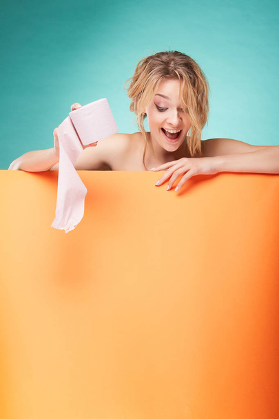 Young cute blonde woman holding roll of toilet paper and looking down with smiling face in studio on turquoise background and orange sheet  - Zdjęcie, obraz