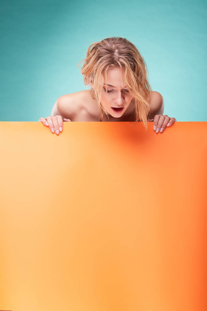 Young surprised blonde woman looking down in studio with turquoise background and orange sheet - Foto, afbeelding