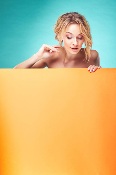 Fiatal aranyos szőke nő nézett le a türkiz háttér és narancssárga lap stúdió - Fotó, kép