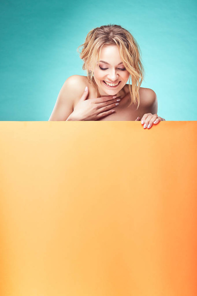 Young cute blonde woman looking down and smiling in studio with turquoise background and orange sheet - 写真・画像