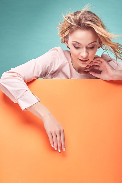 Portrait of young amazing blonde woman looking down in studio with turquoise background and orange sheet - Photo, image