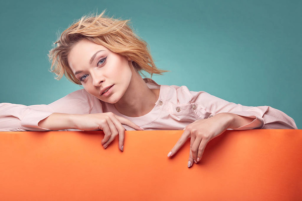 Closeup portrait of young amazing blonde woman with sincere face in studio with turquoise background and orange sheet - Photo, Image