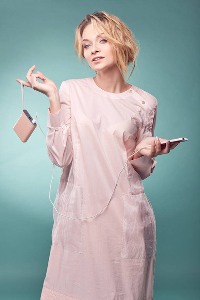 Young romantic blonde woman in light pink dress posing in studio with pink smartphone on turquoise background - Foto, Imagen