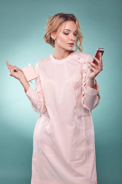 Young romantic blonde woman with wavy hair in light pink dress looking at her pink smartphone in studio on turquoise background - 写真・画像