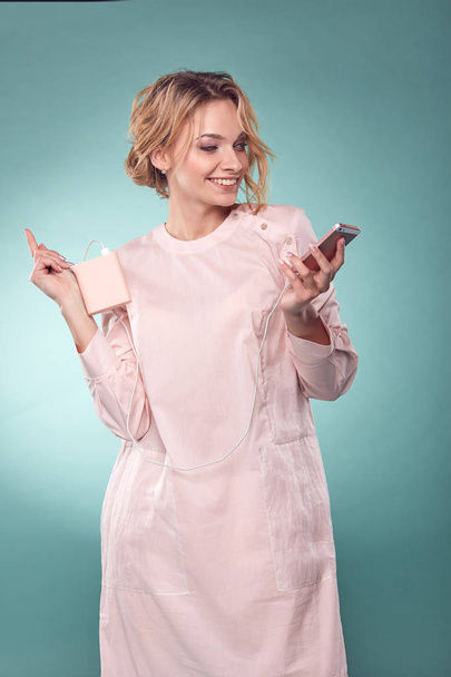 Young romantic smiling blonde woman with wavy hair in light pink dress holding pink smartphone in studio on turquoise background - Foto, Imagen