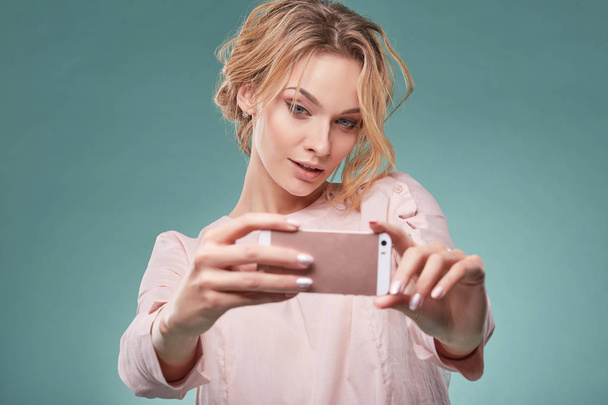 Closeup portrait of young sexy blonde woman with wavy hair in light pink dress looking temptingly at pink smartphone in studio on turquoise background - Zdjęcie, obraz