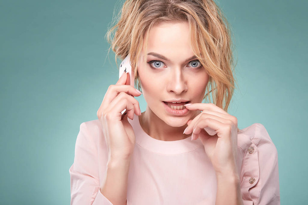 Portrait of young sexy blonde woman with curly hair in light pink dress talking and looking surprised by pink smartphone in studio on turquoise background - Photo, Image