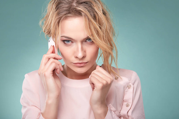 Portrait of young sexy blonde woman with curly hair in light pink dress talking and looking serious by pink smartphone in studio on turquoise background - Photo, image