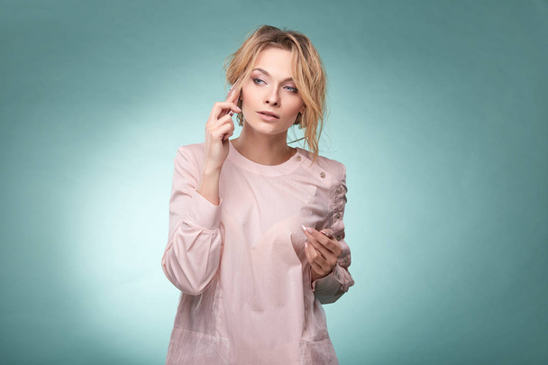 Young pretty blonde woman with wavy hair in light pink dress talking by pink smartphone and looking serious in studio on turquoise background - Photo, Image