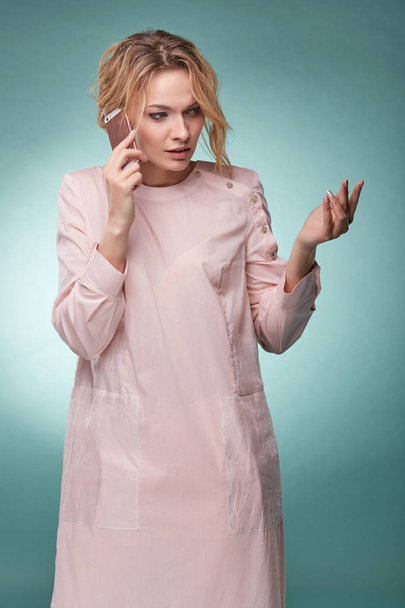 Young pretty blonde woman with wavy hair in light pink dress talking by pink smartphone and looking busy in studio on turquoise background - Valokuva, kuva