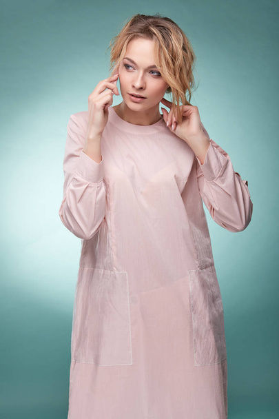 Young pretty blonde woman with wavy hair in light pink dress talking by pink smartphone and looking busy in studio on turquoise background - Fotó, kép