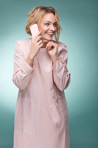 Jonge mooie blonde vrouw met golvend haar in licht roze jurk praten door roze smartphone en lachend in studio op turkooizen achtergrond - Foto, afbeelding