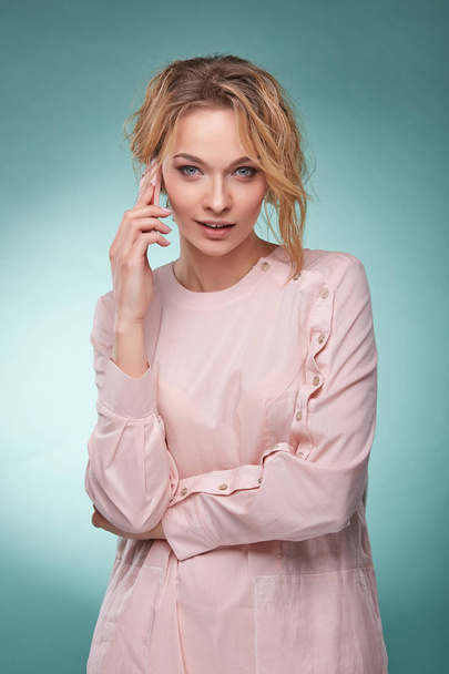 Young beautiful blonde woman with wavy hair in light pink dress talking by pink smartphone and looking temptingly in studio on turquoise background - Foto, Imagen