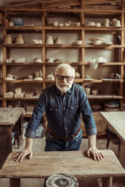 Frontansicht eines älteren Töpfers, der in der Werkstatt auf einem Tisch steht und sich an Regale mit Töpferwaren lehnt - Foto, Bild