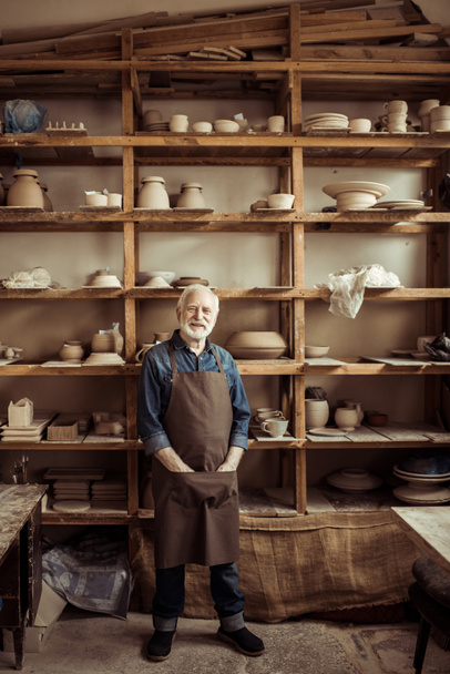 Vista frontal del alfarero senior en delantal de pie contra estantes con productos de cerámica en el taller
 - Foto, Imagen