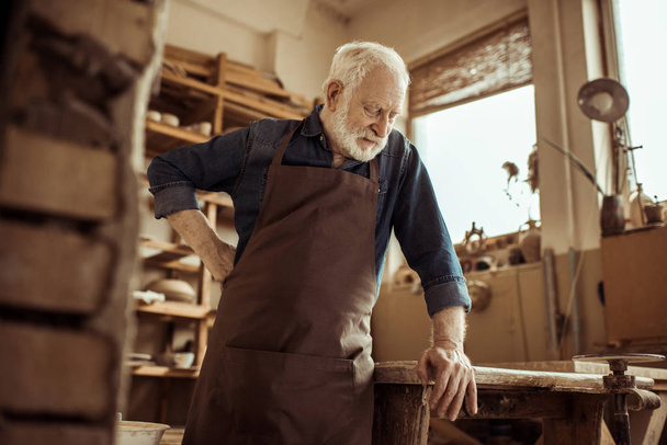 Vista frontal del alfarero senior en delantal de pie y apoyado en la mesa contra los estantes con productos de cerámica en el taller
 - Foto, Imagen