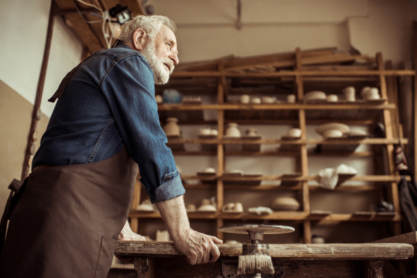 Seitenansicht des Senior-Töpfers in Schürze stehend und auf einem Tisch gegen Regale mit Töpferwaren in der Werkstatt gelehnt - Foto, Bild