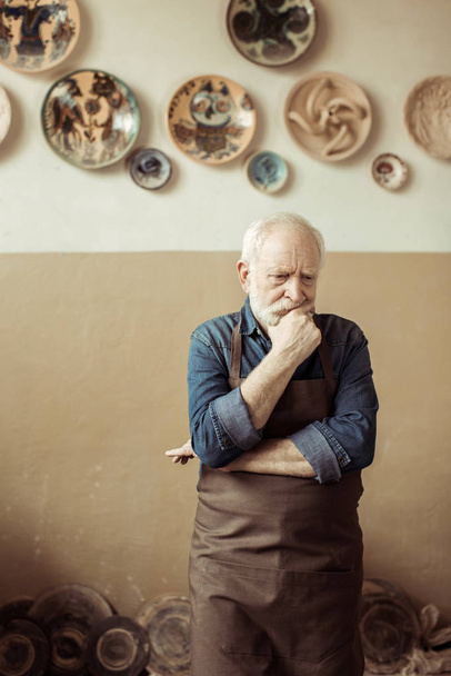 Alfarero senior en delantal pensando y de pie contra la pared con artículos de cerámica colgantes
 - Foto, Imagen